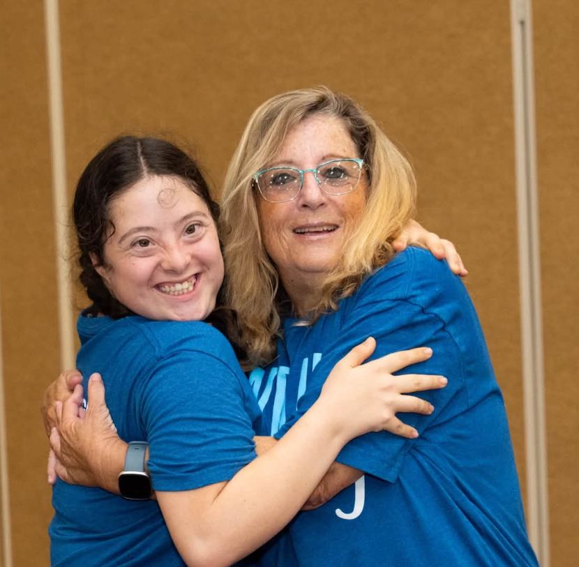 A young woman and a volunteer in matching blue shirts hugging and smiling