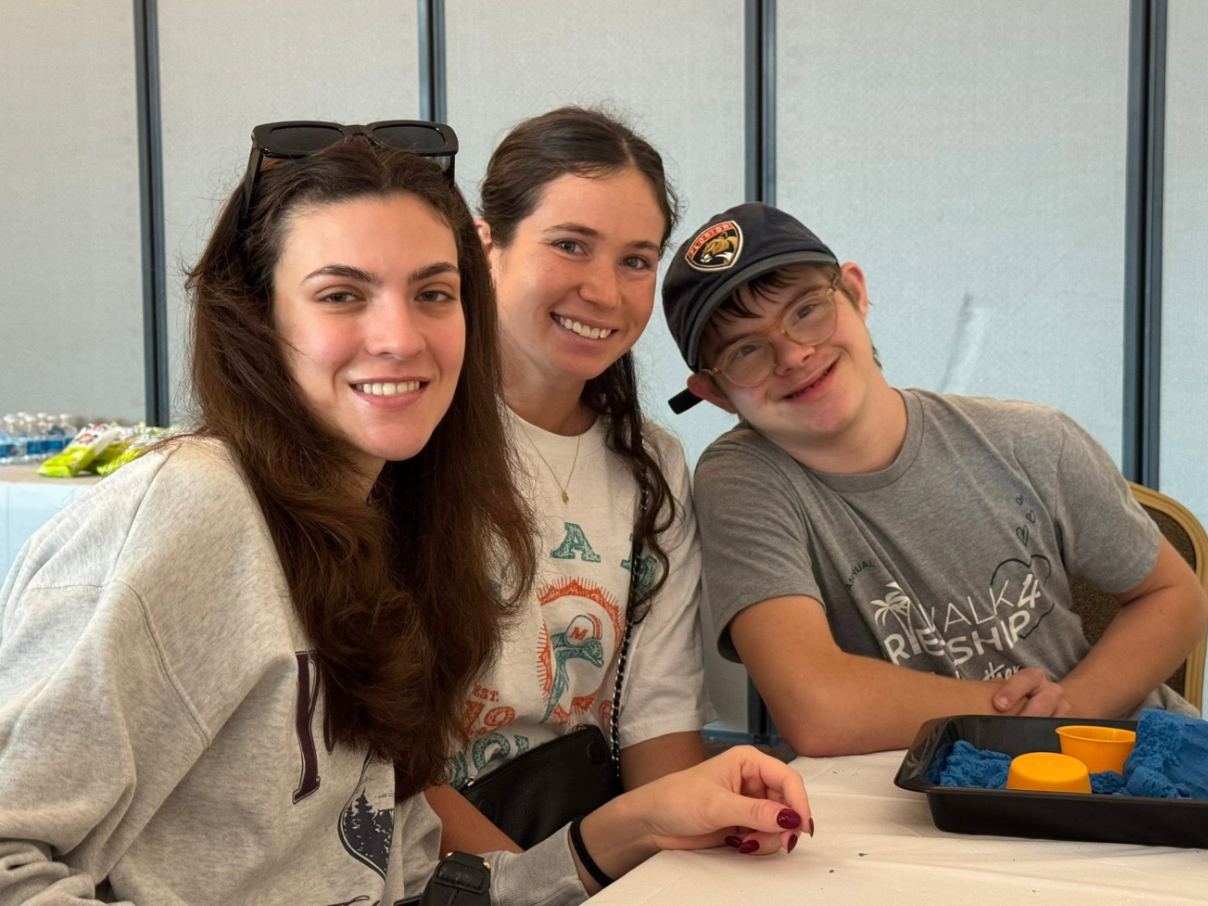 Two smiling volunteers sitting beside a participant in a baseball cap at a Sunday Circle table