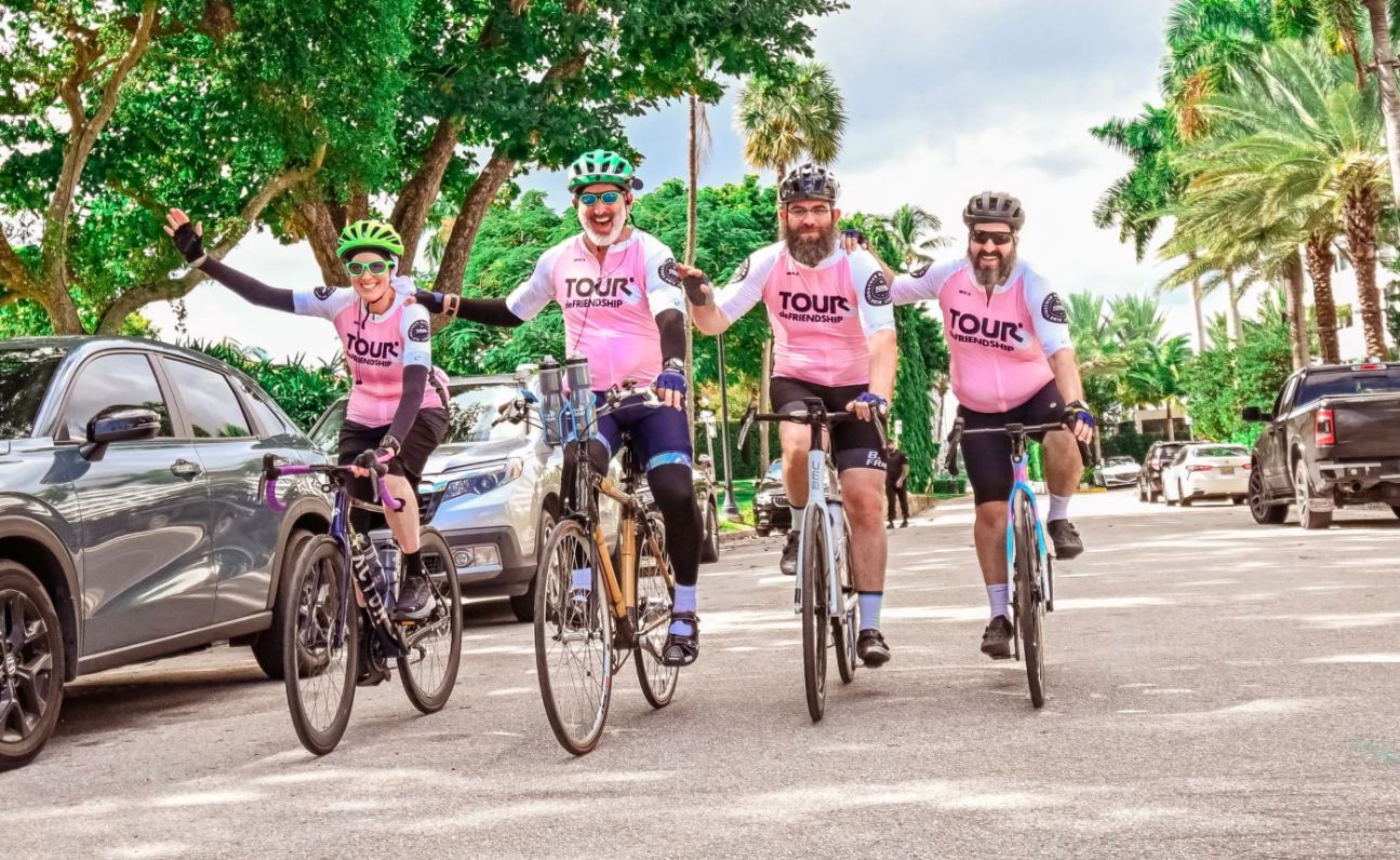 Four riders in pink Tour de Friendship jerseys riding toward the camera, arms out