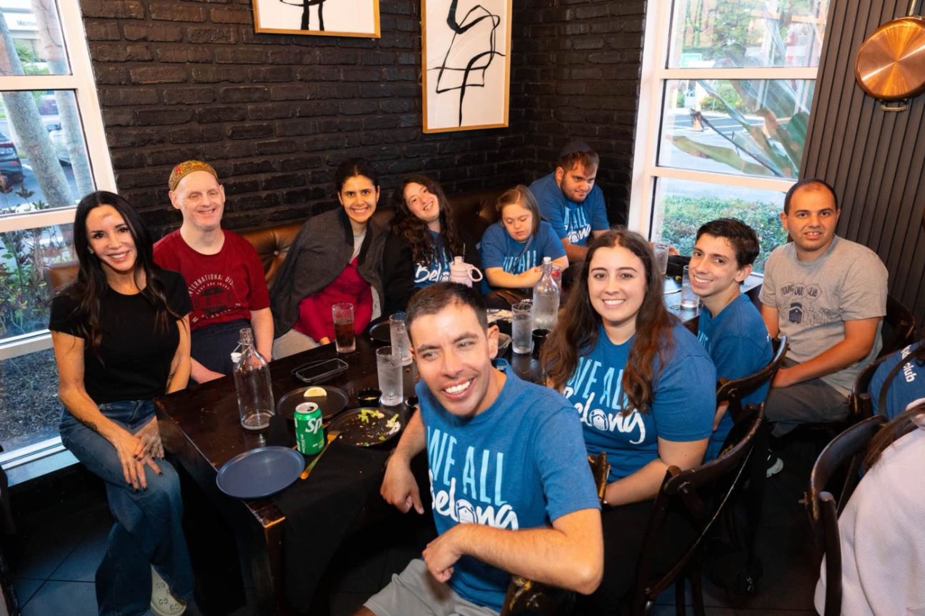 Young adults in We All Belong shirts around a restaurant table, smiling at the camera